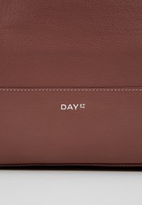 Ruskea nahkalaukku, jossa on nyppyinen pinta, hienovarainen ommelviiva ja valkoinen painettu "DAY ET" -logo.