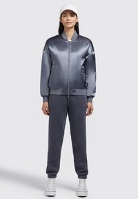 Jachetă bomber din satin, de culoare gri-albastru, cu manșete și tiv din tricot. Asortată cu pantaloni de trening gri. Tenisi albi cu glezna înaltă completează ținuta.