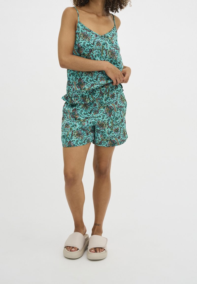 Femme portant un haut sans manches à fleurs turquoise et un short assorti, avec des sandales beige à plateforme, debout devant un fond uni.