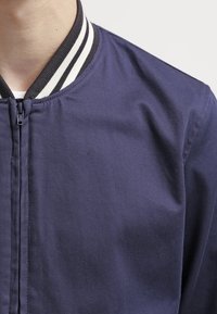 Veste aviateur bleu marine avec col côtelé rayé bicolore, fermeture éclair et texture de tissu lisse ; vue rapprochée de la partie supérieure avant.