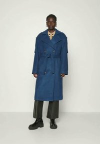 Blauwe, lange wollen jas met belted taille, grote revers en dubbelzijdige knopen. Gestyled over zwarte, wijde broekspijpen en chunky laarzen.