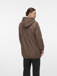 Moteris su surištais plaukais, vilkinti rudą gobtuvą ir juodas kelnes, stovi dešinėje pusėje prieš paprastą fono.