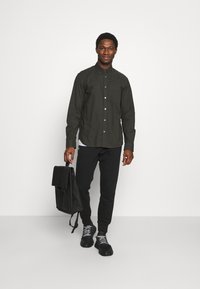 Camisa verde-escura de botões, calças de fato pretas e ténis pretos. Cabelo curto, segurando uma mochila preta com aba. Fundo branco.