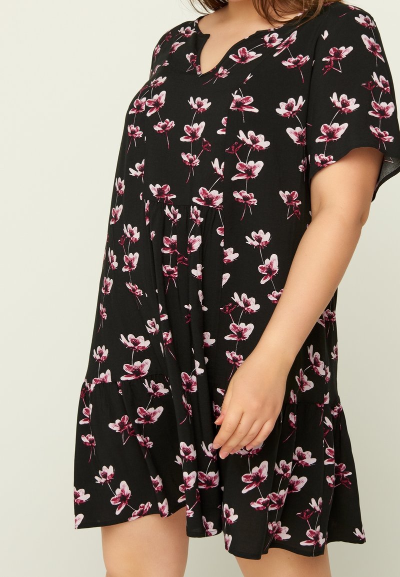 Svart kortärmad klänning med rosa och vita blommotiv, med en nederdel med skärning och volanger, bärs av en person med handen vila på kjolen.