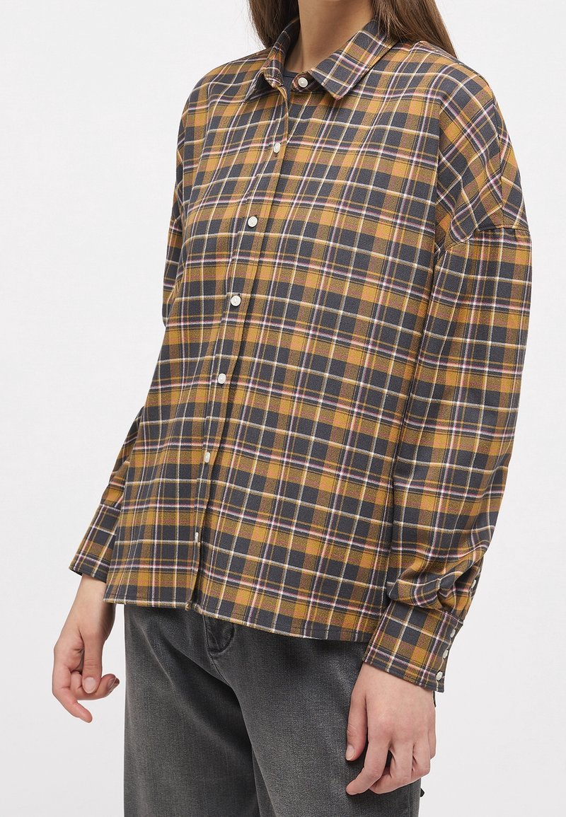 Femme portant une chemise à carreaux jaune, noire et blanche, à manches longues, avec un pantalon gris foncé.