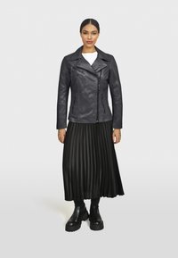 Čierna kožená motorkárska bunda s šikmým zipsom, skombinovaná s plisovanou čiernou sukňou a robustnými čiernymi čižmami. Obsahuje hladkú textúru a štruktúrovaný dizajn.