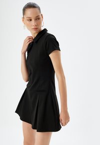 Vestido polo negro con mangas cortas, corsé ajustado y falda acampanada. Presenta un cuello y bolsillos delanteros con un sutil detalle de costura.