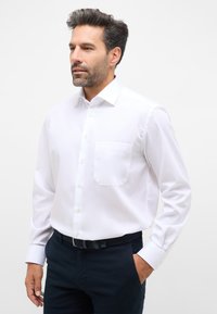 Camicia bianca a maniche lunghe con colletto a punta, taschino sul petto e chiusura con bottoni. Indossata con pantaloni blu e una cintura strutturata.