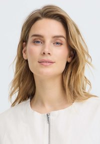 Jeune femme aux cheveux blonds clairs et aux yeux bleus, portant un haut blanc à fermeture éclair, regardant doucement la caméra sur un fond uni.