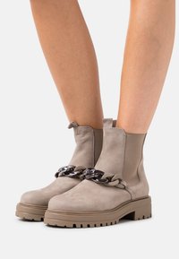 Botas de tobillo de ante beige con suela de goma gruesa, que presentan una cadena decorativa en negro y beige en la parte delantera y paneles laterales elásticos.
