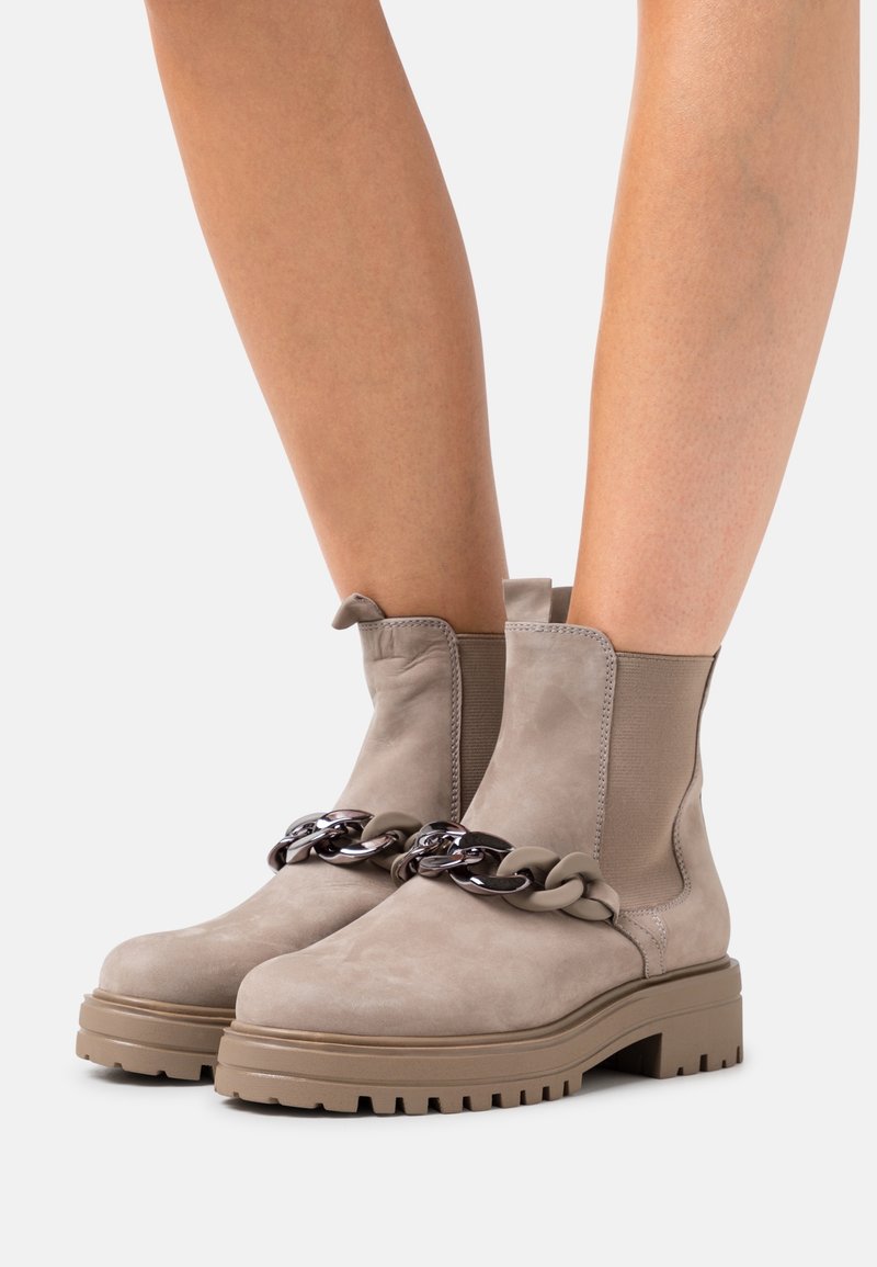 Botas de tobillo de ante beige con suela de goma gruesa, que presentan una cadena decorativa en negro y beige en la parte delantera y paneles laterales elásticos.