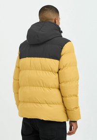 Gelbe und schwarze Steppjacke mit Kapuze, ausgestattet mit einem diamantgeprägten Design und einer glatten Textur, sichtbaren Nähten und einem kontrastierenden Farbblock.