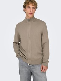 Pull beige à fermeture éclair avec un col haut et des poignets côtelés. Texture douce avec une coupe ajustée, porté sur un jean gris clair.