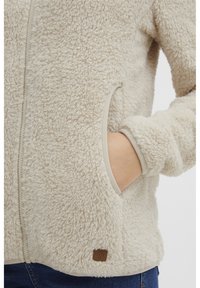 Beige fleecejakke med en teksturert overflate, med glidelås og sidelomme. Akkompagnert av et lite brunt logo-lerret.