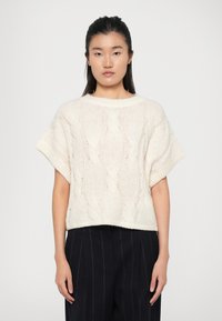 Cremfarbener Kabelstrickpullover mit lockerer Passform, kurzen Ärmeln und geripptem Saum. Getragen über dunklen gestreiften Hosen.