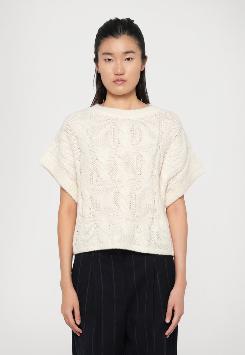 Cremfarbener Kabelstrickpullover mit lockerer Passform, kurzen Ärmeln und geripptem Saum. Getragen über dunklen gestreiften Hosen.