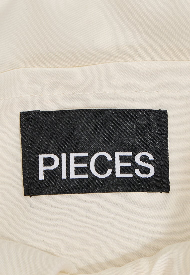Etiqueta con la palabra "PIECES" en un elegante tipo de letra blanca sobre un fondo negro, unida a una tela de color crema con una textura suave.