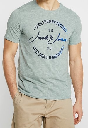 Homme portant un t-shirt à manches courtes vert clair avec le logo "Jack & Jones" et un texte circulaire, associé à un pantalon beige.