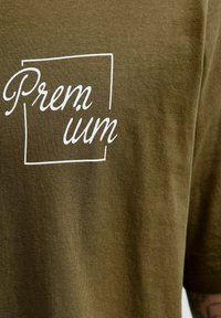 Olivově zelená látka s bílým kurzívním textem "Premium" částečně uzavřeným v neúplném čtvercovém obrysu.