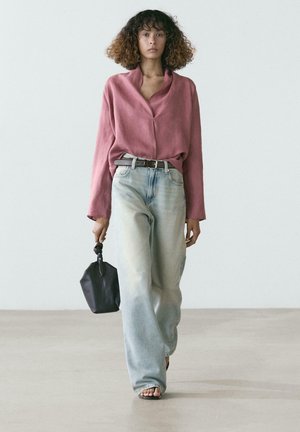 Femme aux cheveux bouclés portant un chemisier rose ample, un jean large clair, tenant un petit sac à main noir, marchant sur un sol clair devant un mur uni.