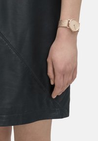 Rundgesichtige Uhr mit einem Gehäuse aus Roségold und einem Metallnetzarmband, kombiniert mit einem schwarzen Ledermantel mit diagonalen Nähten.