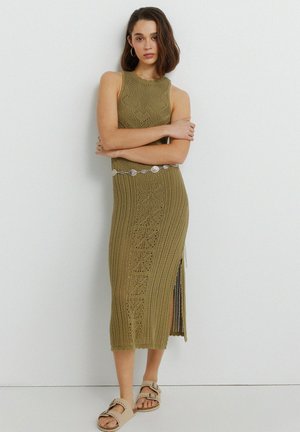 Donna in piedi con le braccia incrociate, che indossa un vestito senza maniche in maglia verde oliva con spacco laterale e sandali beige con fibbia.