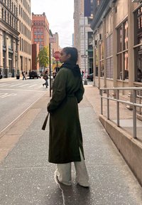 Dlhý olivovo zelený kabát s kapucňou, nosený cez svetlošedé nohavice s širokými nohavicami. Ulica s mestskými budovami a chodcami v pozadí.