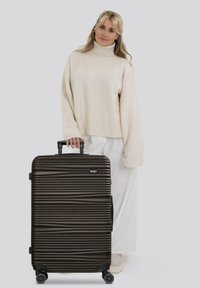 Vrouw in een beige coltrui en witte broeken, staand met een grote donkere harde koffer met horizontale textuurlijnen.