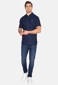 Marineblaue Kurzarm-Hemd mit Knöpfen und subtiler Logo auf der Brust, kombiniert mit dunkelblauen Skinny-Jeans und weißen Sneakers.