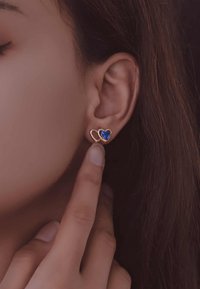Pendientes en tono dorado con formas de corazones entrelazados y una gema azul en el centro, exhibidos en una mano cerca de una oreja.