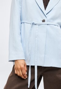 Personne portant une veste bleu clair ceinturée par-dessus une chemise à pois marron foncé et un pantalon marron foncé, la main reposant le long du corps.