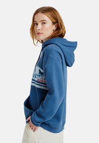 Sudadera azul hecha de tela suave, con un diseño gráfico en el frente que incluye montañas y olas, y un bolsillo tipo canguro.
