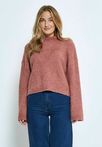 Pullover aus weichem rosafarbenem Strickmaterial, mit einem hohen Kragen und weiten Ärmeln, mit einem verkürzten Saum über dunkelbblauen Jeans.