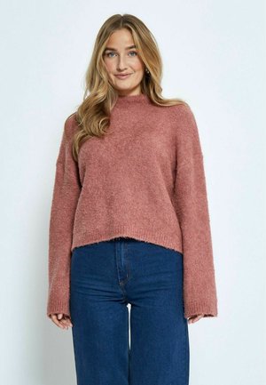 Trui gemaakt van zacht roze breimateriaal, met een hoge hals en wijde mouwen, met een cropped zoom bovenop donkerblauwe jeans.