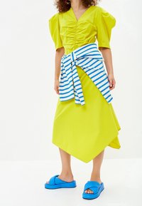 Limetkově zelené šaty s nabíranými rukávy a nařaseným předním dílem, doplněné modro-bílým pruhovaným opaskem kolem pasu a masivními modrými sandály.