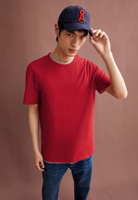 Rotes Baumwoll-T-Shirt mit kurzen Ärmeln und Rundhalsausschnitt, kombiniert mit dunkelblauen Jeans und einer marineblauen Kappe mit rotem Logo-Akzent.