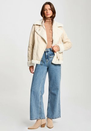 Giubbotto beige in shearling con colletto a punta, chiusura con zip e fodera crema, abbinato a jeans blu a vita alta e stivaletti beige.