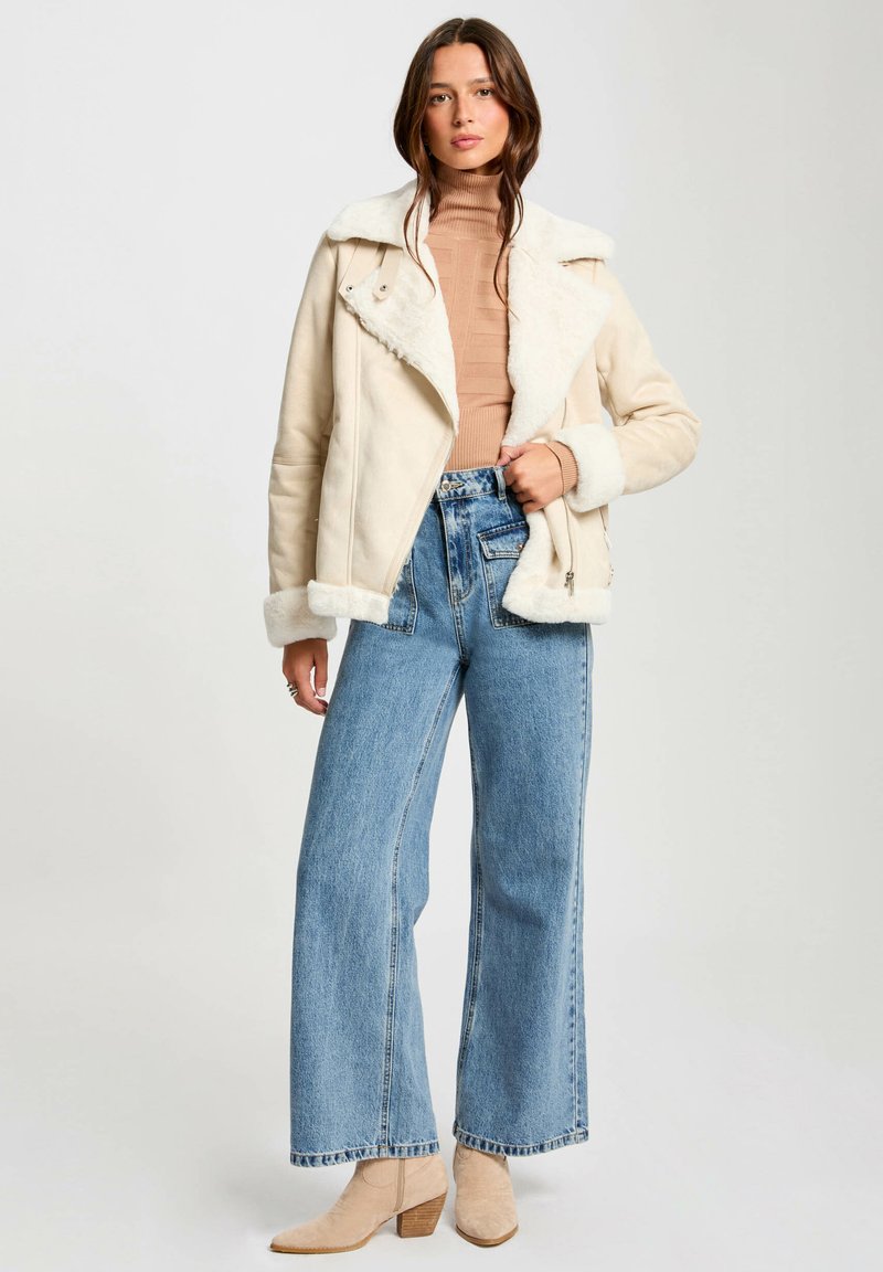 Veste en shearling beige avec un col à revers, fermeture éclair à l'avant et doublure crème, associée à un jean en denim bleu taille haute et des bottines beige.