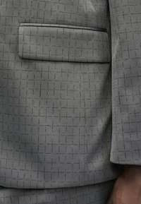 Hallikasbleiseri kangas, millel on tekstuuriga muster, kus lõikuvad jooned. Vasakul küljel on welt-tasku.