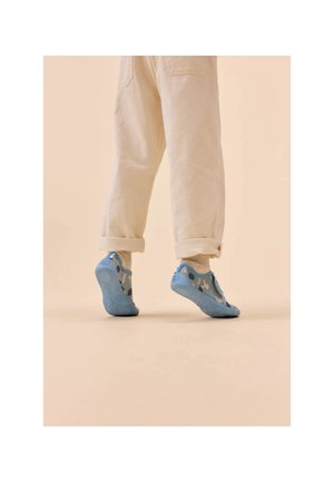 Chaussures bleues avec une semelle texturée et un motif camouflage, portées avec un pantalon crème retroussé et des chaussettes claires, sur un fond neutre.