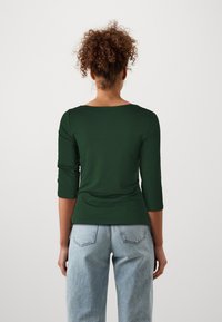 Green, fitted top with three-quarter sleeves and a wide neckline. Worn with light wash, high-waisted jeans. Smooth texture.