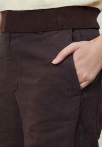 Brązowe bawełniane spodnie z ryflem w pasie, wyposażone w boczne kieszenie i gładką fakturę. Ręka umieszczona w kieszeni, ukazująca detal tkaniny.