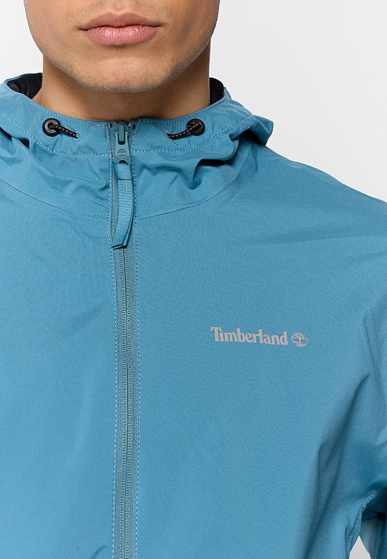 Leichte blaue Jacke mit Kapuze, ausgestattet mit einem Frontreißverschluss, verstellbarem Kordelzug und weißen Timberland-Logo auf der linken Brust.