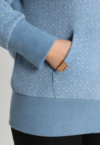 Mano dentro del bolsillo delantero de un sudadera azul con lunares, con puños y dobladillo de canalé, mostrando una pequeña etiqueta de tela marrón de la marca en el borde del bolsillo.