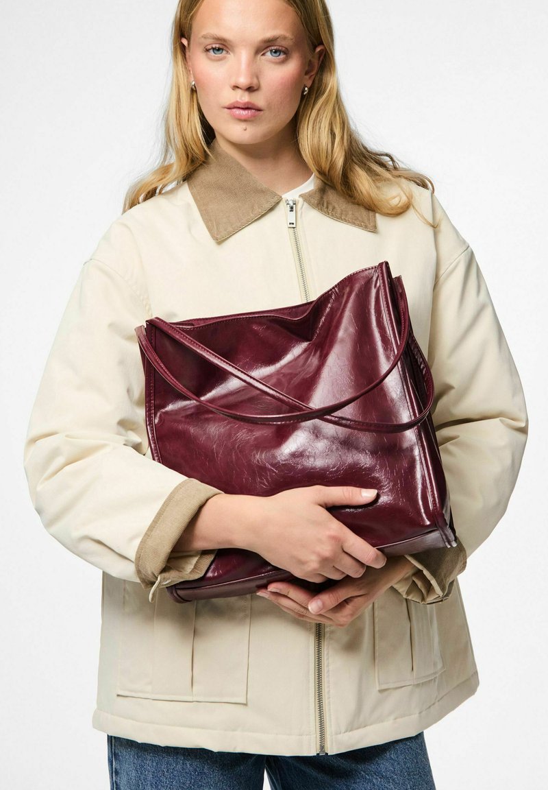 Donna con i capelli biondi lunghi che indossa una giacca beige tiene una grande borsa in pelle lucida rossa scura contro uno sfondo bianco.