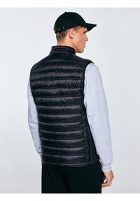 Férfi fekete steppelt ujjatlan dzsekit visel szürke pulóver felett és fekete sapkát, oldalra néz, egyszerű háttér előtt.