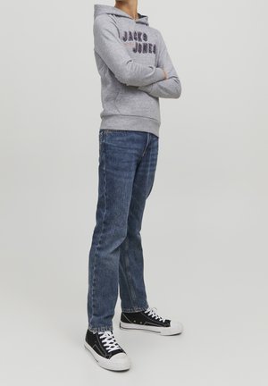 Person stående med armene krydset iført en grå Jack & Jones hættetrøje, blå jeans og sorte høje sneakers på en ensfarvet baggrund.