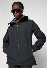 Woman wearing a black Didriksons hooded jacket with hands in pockets and large black sunglasses against a plain background.