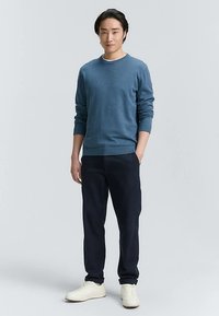 Blå crewneck-tröja, mörka byxor och vita sneakers. Tröjan har ribbade mudd; byxorna är raka i benen. Enkel, avslappnad outfit.