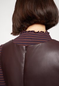 Robe en cuir marron avec un dos texturé, dotée d'un col à volants et d'une fermeture éclair, combinée avec un motif plaid foncé sur la partie supérieure.